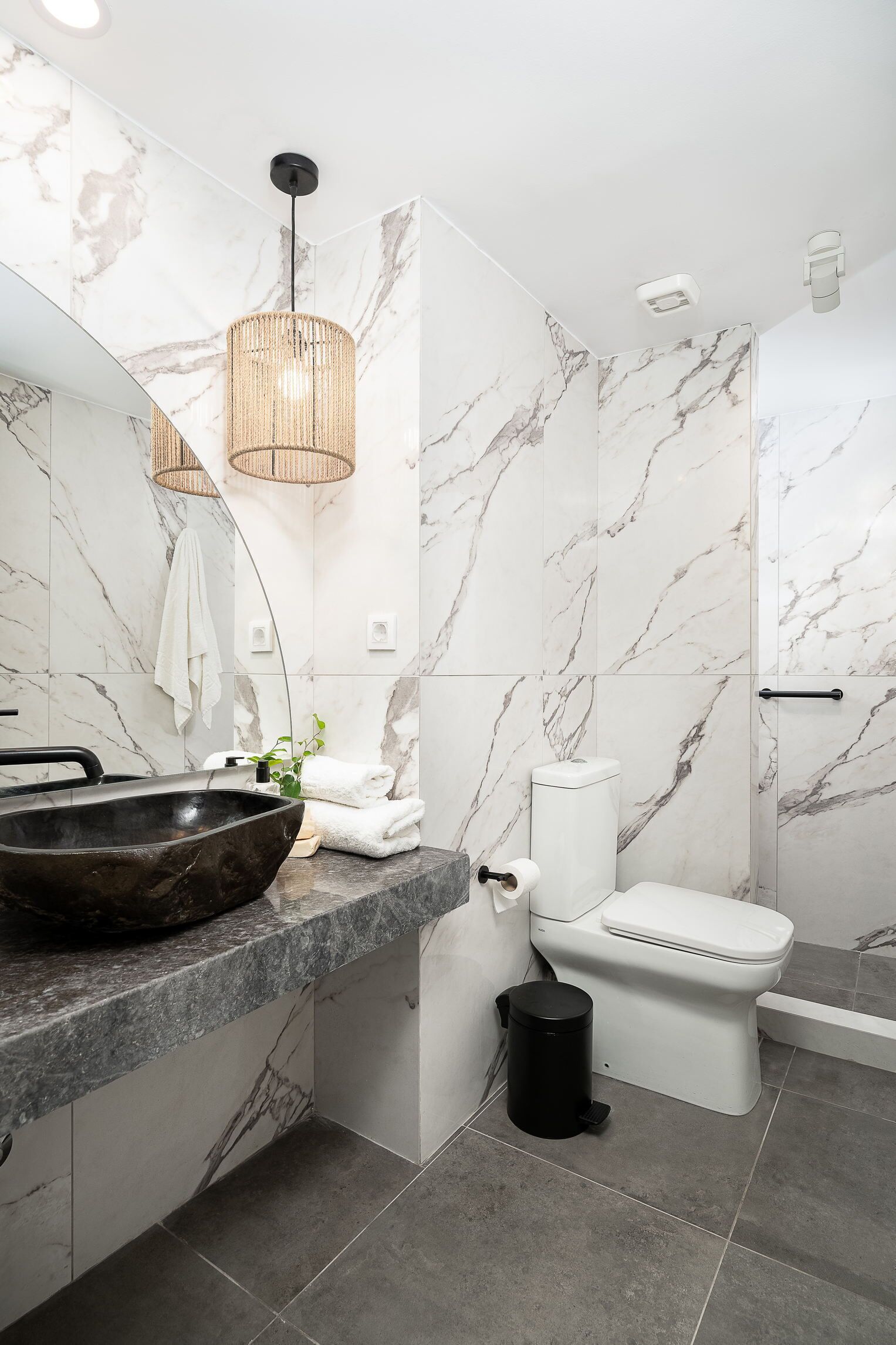 Une salle de bain moderne avec un pare-baignoire en marbre, un lavabo sur pied noir, des toilettes et une poubelle noire.
