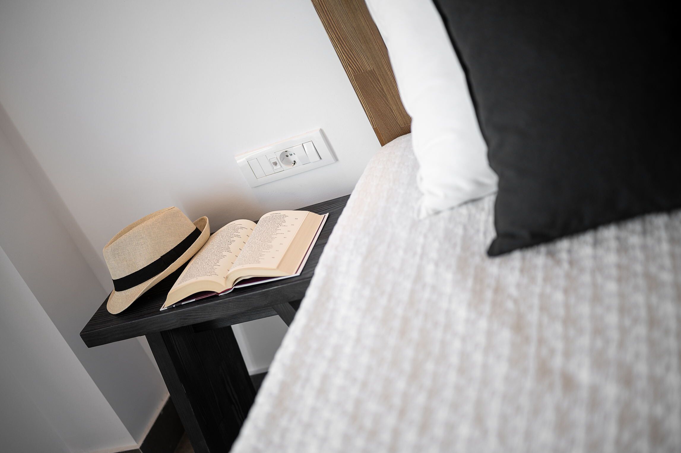 Un lit bien fait, avec une couette blanche et un oreiller noir, une petite table sur laquelle se trouvent un chapeau et un livre ouvert, et une prise de courant au mur.