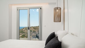 Een slaapkamer met een bed, twee kussens, een hanglamp en een schuifdeur die leidt naar een balkon met uitzicht op een rotsachtig landschap.