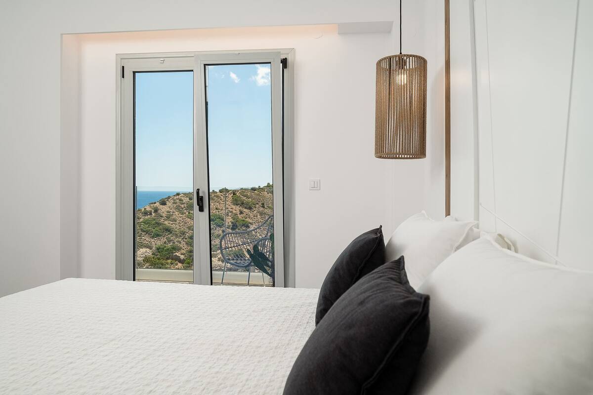 Un dormitori amb un llit, dos almoinos, una làmpada penjant i una porta corredissa de vidre que dona a un balcó amb vista a un paisatge rocos.