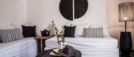 A modern living room with a white sofa, black and white patterned pillows, a round black and white wall art piece, a small wooden side table with a book and a teapot, a black coffee table with a hat and book, and a tall black floor lamp.