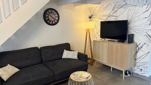 A modern living room with a dark sofa, a small round table with magazines, a TV on a wooden cabinet, and a wall clock. - Maison au Cœur de La Palmyre (Les Mathes)