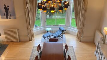A dining room with a wooden table and chairs, a chandelier, and a view of the garden through large windows.