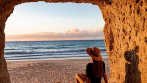 Une personne est assise sur une plage de sable, et observe l’océan à travers une structure rocheuse qui ressemble à une grotte.