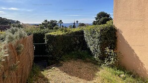 A courtyard with a view of the sea, surrounded by greenery and a building with a textured wall.