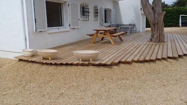 A white building with a sloped roof, a wooden deck with a picnic table and benches, and a tree in the background. - Maison de Vacances à 15 Km de Saint-jean de Mont (Sallertaine)