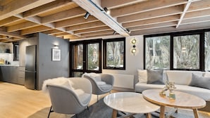A modern living room with grey sofas, a round coffee table, and a kitchen area with a fridge and cabinets. Large windows offer views of trees and a snowy landscape. - Alpine Uralla - Dinner Plain (Dinner Plain)