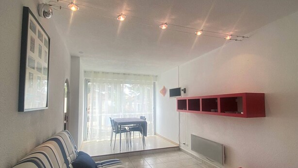 Una sala de estar con un sofá a rayas, un banco rojo, un televisor montado en la pared y un comedor con mesa y sillas.