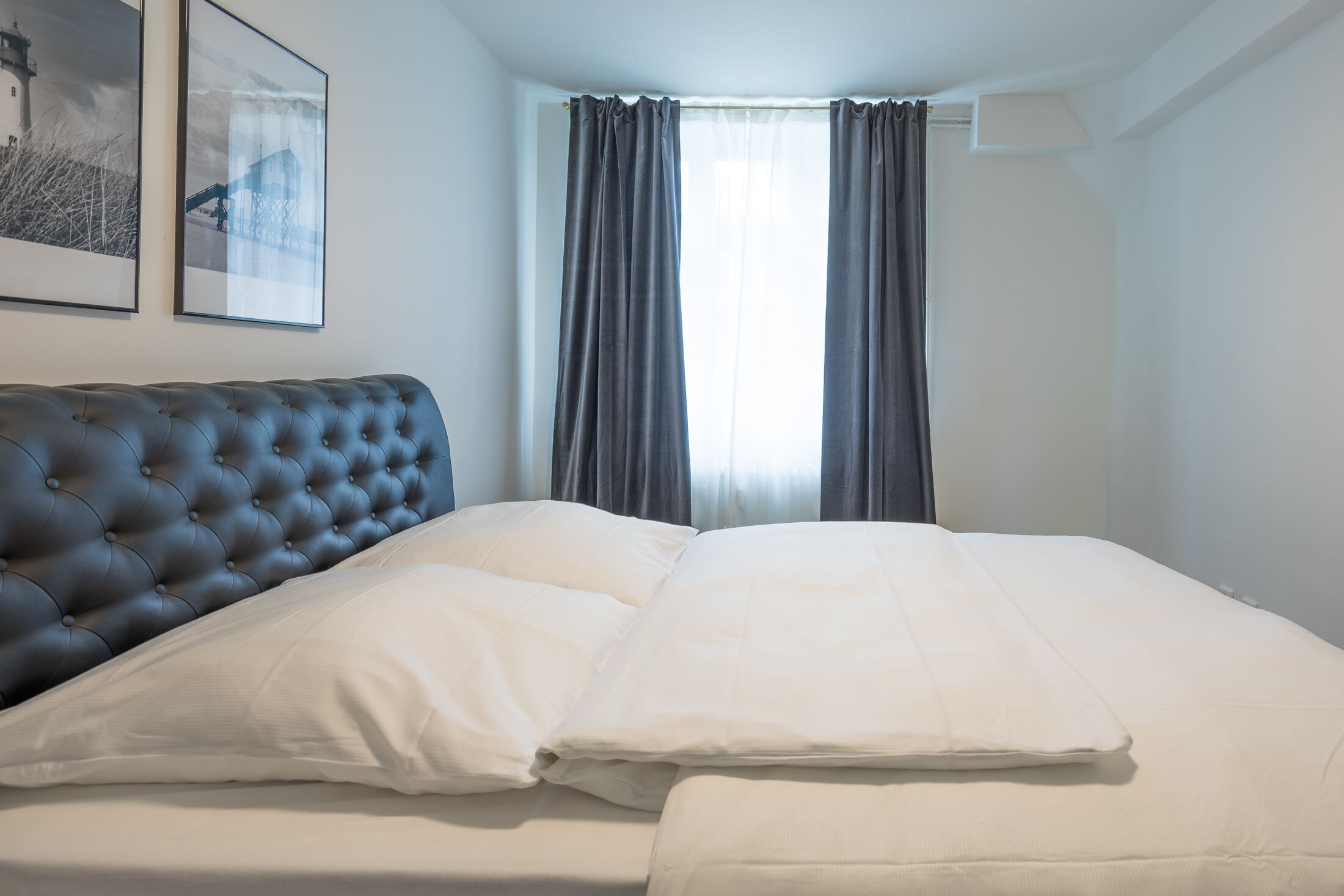 Een keurig opgemaakt bed met witte lakens en een geborduurde hoofdbord, een raam met gordijnen, en twee ingelijste foto's aan de muur.