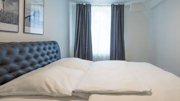 Een keurig opgemaakt bed met witte lakens en een geborduurde hoofdbord, een raam met gordijnen, en twee ingelijste foto's aan de muur.