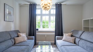 A modern living room with two grey sofas, a glass coffee table, a radiator, and a window with curtains.
