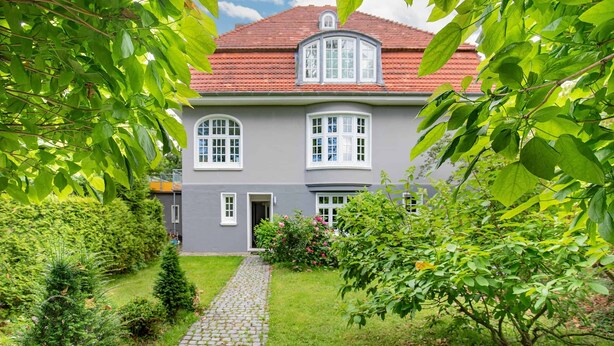 A two-story house with a red-tiled roof, a grey facade, and a cobblestone pathway leading to the entrance.