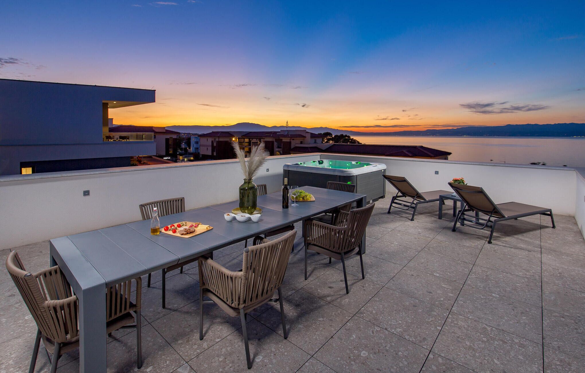 Eine Dachterrasse mit Esstisch, Stühlen, einem Whirlpool und Blick auf den Sonnenuntergang über dem Wasser.