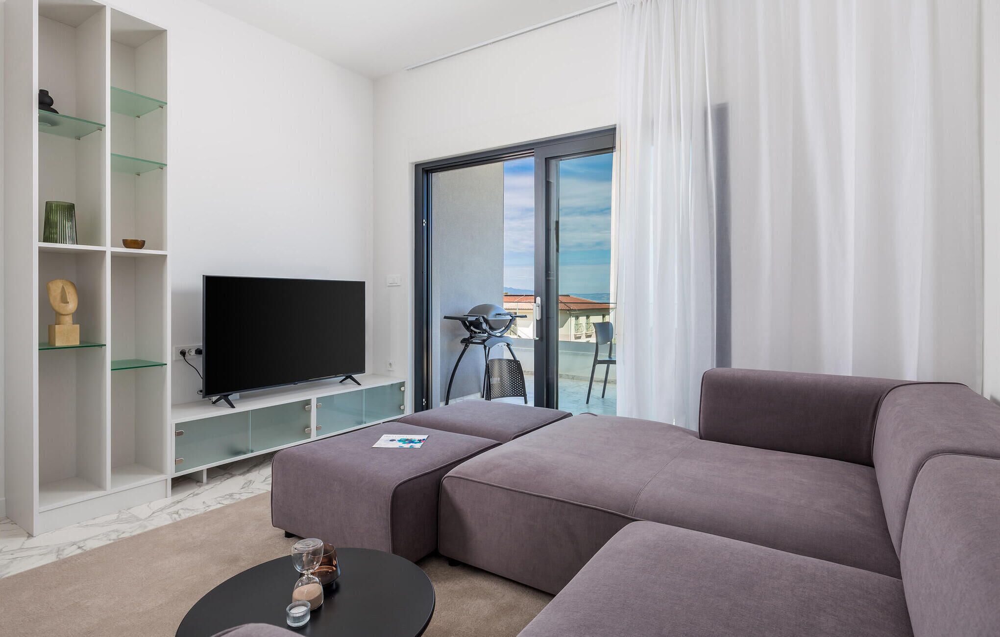Ett modernt vardagsrum med en stor platt-TV, en grå hörnsoffa, ett soffbord i glas och en balkong med utsikt.