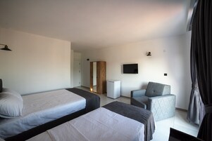 Una habitación de hotel con dos camas individuales, un sillón gris, un armario pequeño blanco y un televisor de pantalla plana montado en la pared.