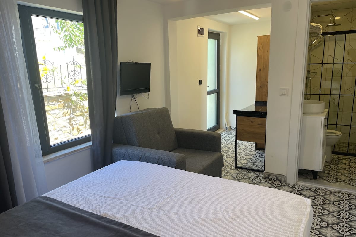 A hotel room with a bed, a grey armchair, a TV mounted on the wall, and a bathroom visible through an open door.