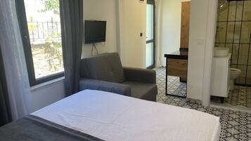 A hotel room with a bed, a grey armchair, a TV mounted on the wall, and a bathroom visible through an open door.