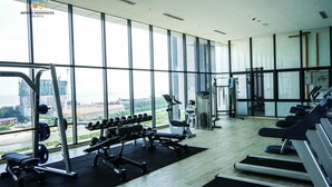 Un gimnasio con equipo de levantamiento de pesas y cintas de correr, ventanas amplias y vistas a un paisaje urbano.