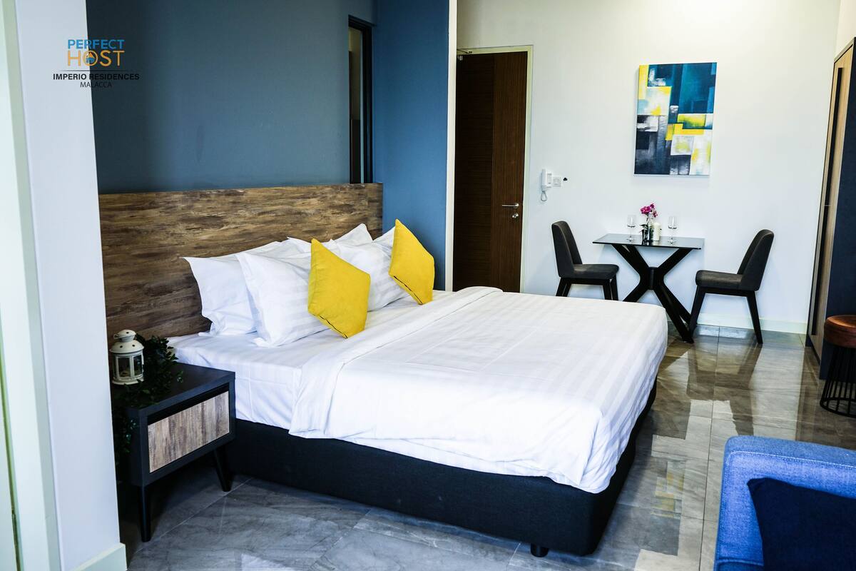 Quarto moderno com uma cama grande, uma mesinha de cabeceira, uma escrivaninha com duas cadeiras e um quadro na parede.
