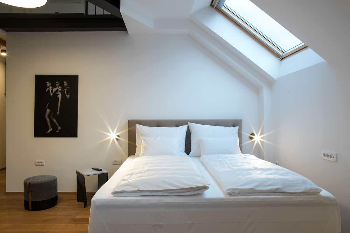 Une chambre avec un puits de lumière, un lit avec du linge de lit blanc, une table de chevet et un petit pouf rond.