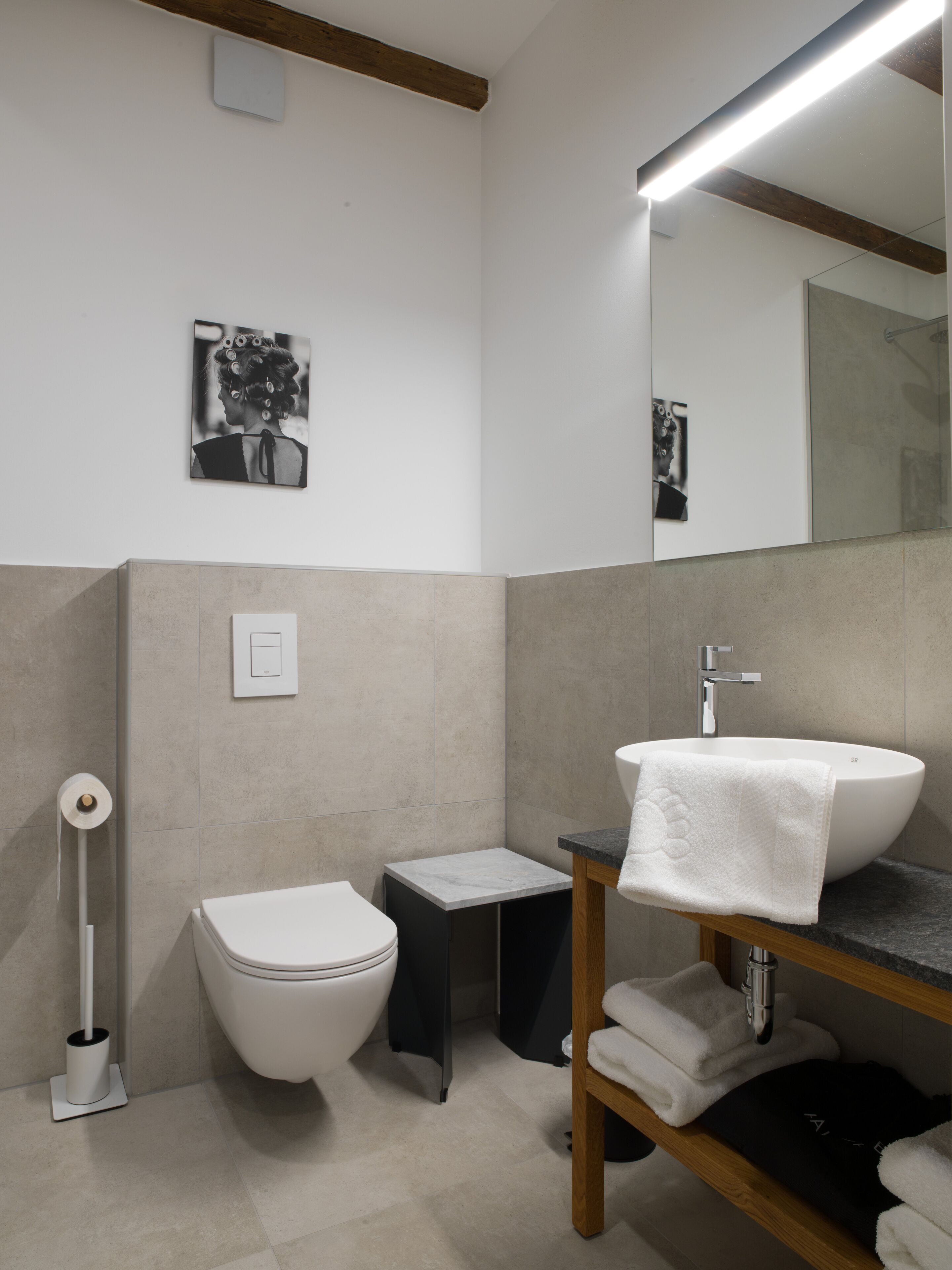 Ein modernes Badezimmer mit Toilette in Weiß, einem kleinen schwarzen Schrank, einem Waschbecken mit Holzablage und einem Spiegel darüber.