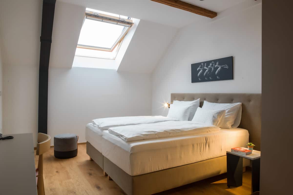 Une chambre à coucher avec un lit, une petite table sur laquelle repose un livre, une œuvre d’art accrochée au mur et une lucarne.