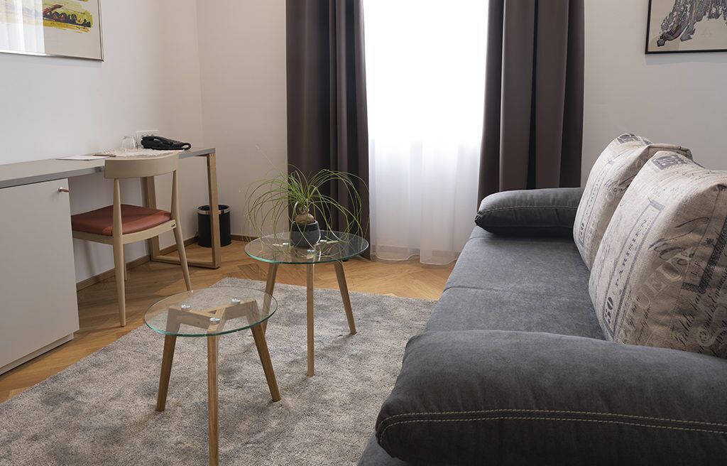 A modern living room with a grey sofa, a glass coffee table, a wooden chair, and a small table with a plant.