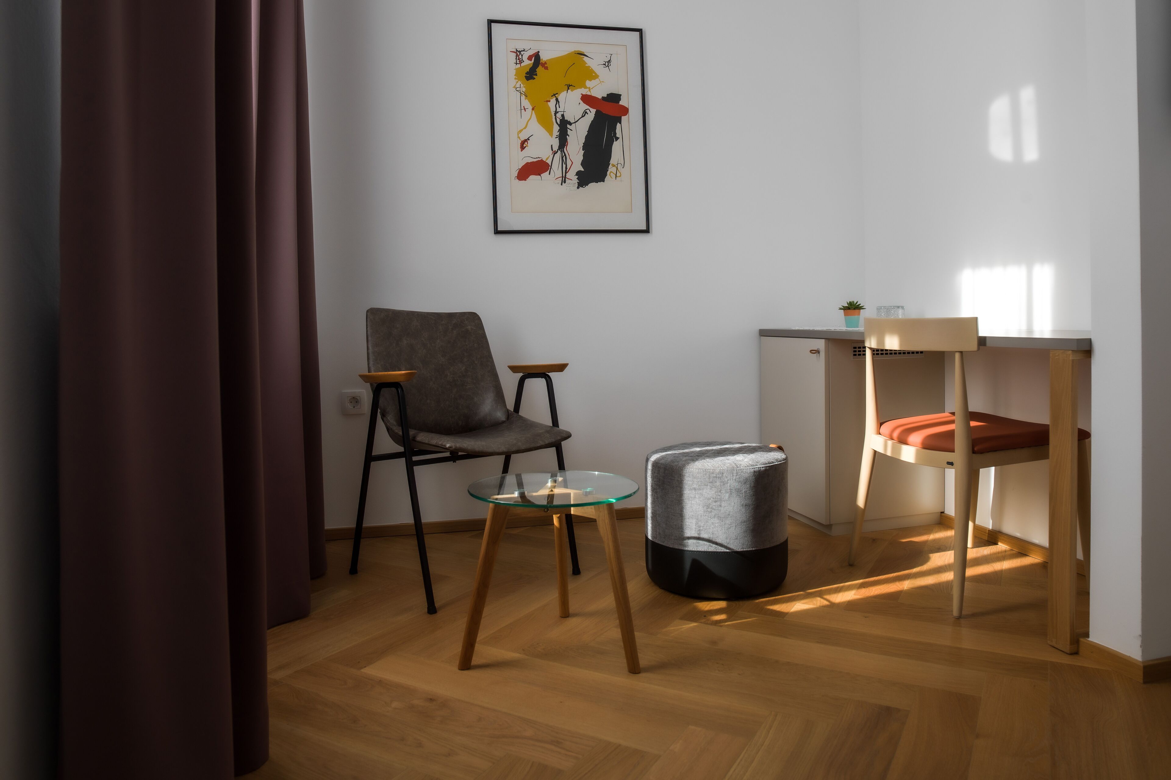 A modern room with a grey chair, a glass table, a small ottoman, and a framed abstract painting on the wall.