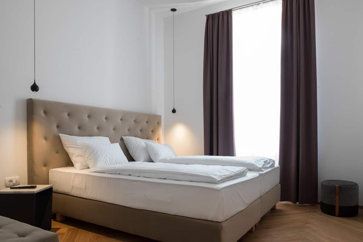 Une chambre moderne avec une tête de lit capitonnée, un grand lit avec du linge de lit blanc, une table de chevet et un pouf rond.