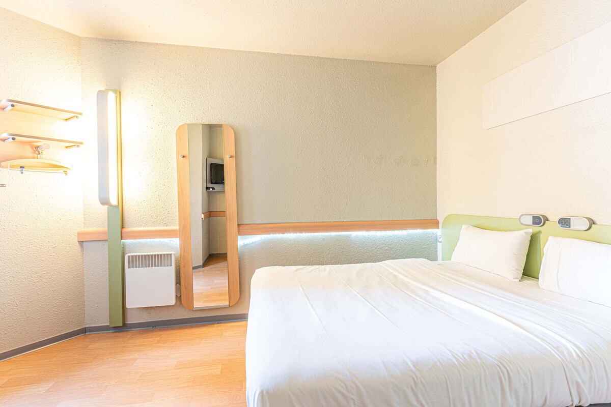 A hotel room with a large bed, a mirror, a heater, and a wall-mounted shelf.