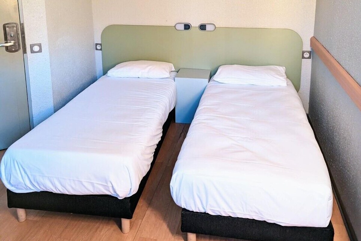 A small, single-room hotel room with two single beds, a small table, and a wall-mounted light fixture.
