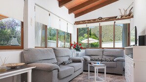 A living room with two grey sofas, a small white table, and a white cabinet. Large windows offer views of greenery and a white fence. - Villa Luna with private pool near the beach for 6 persons, AC, WiFi (Alcudia)