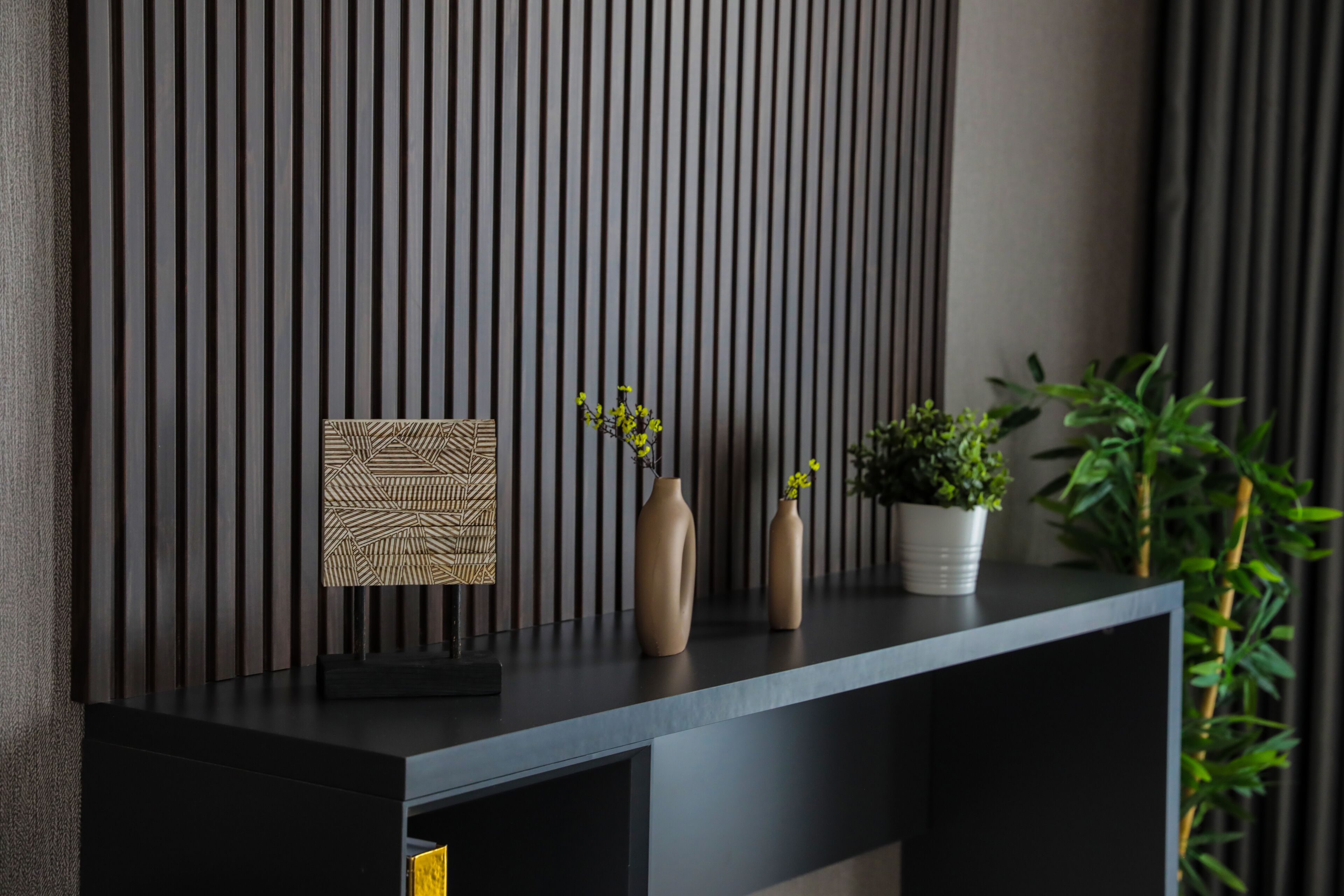 A modern, dark-toned console table with a geometric patterned artwork, two vases with yellow flowers, and a potted plant.