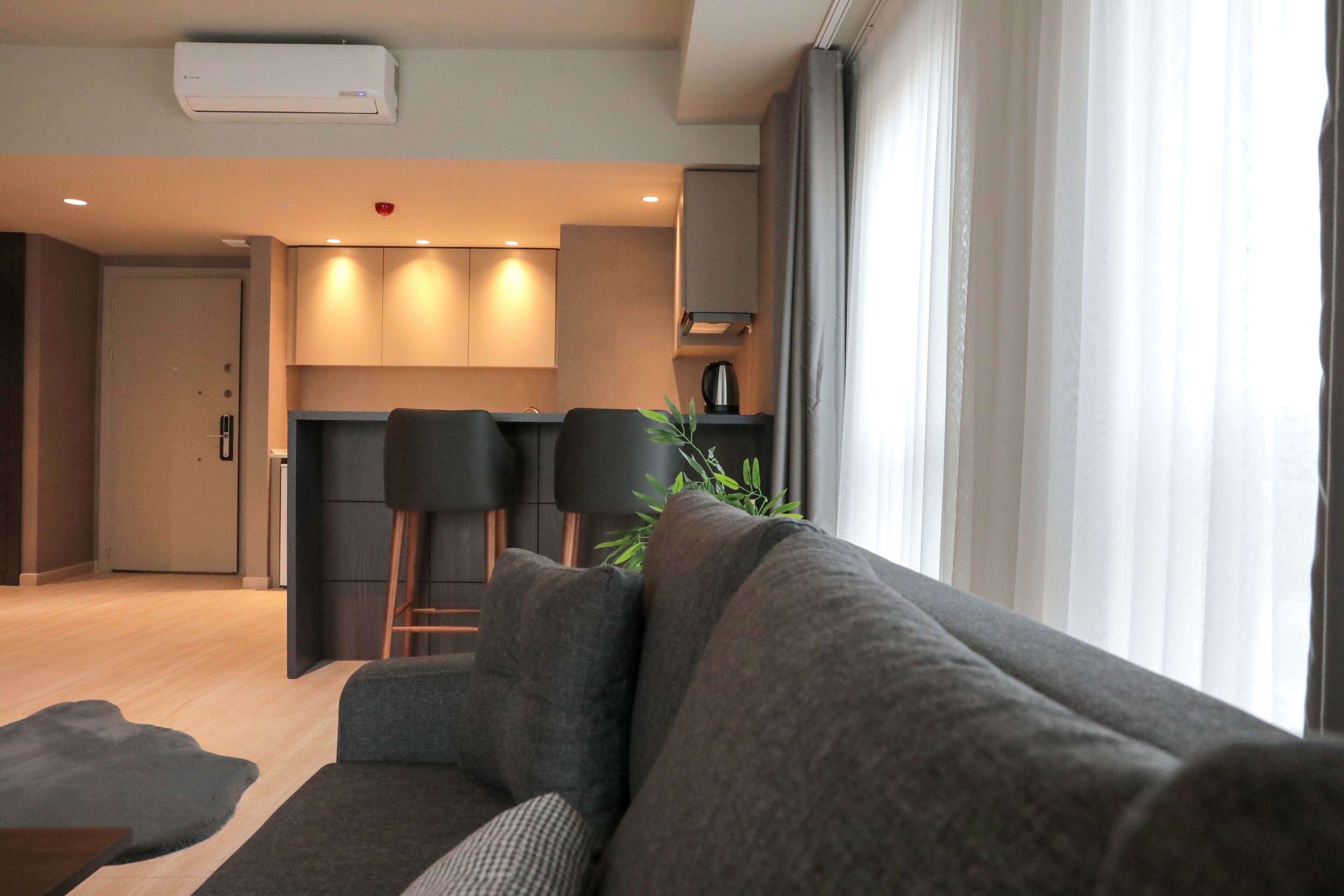 A modern living room with a grey sofa, a coffee table, and a view of a kitchen area with two bar stools and a plant.