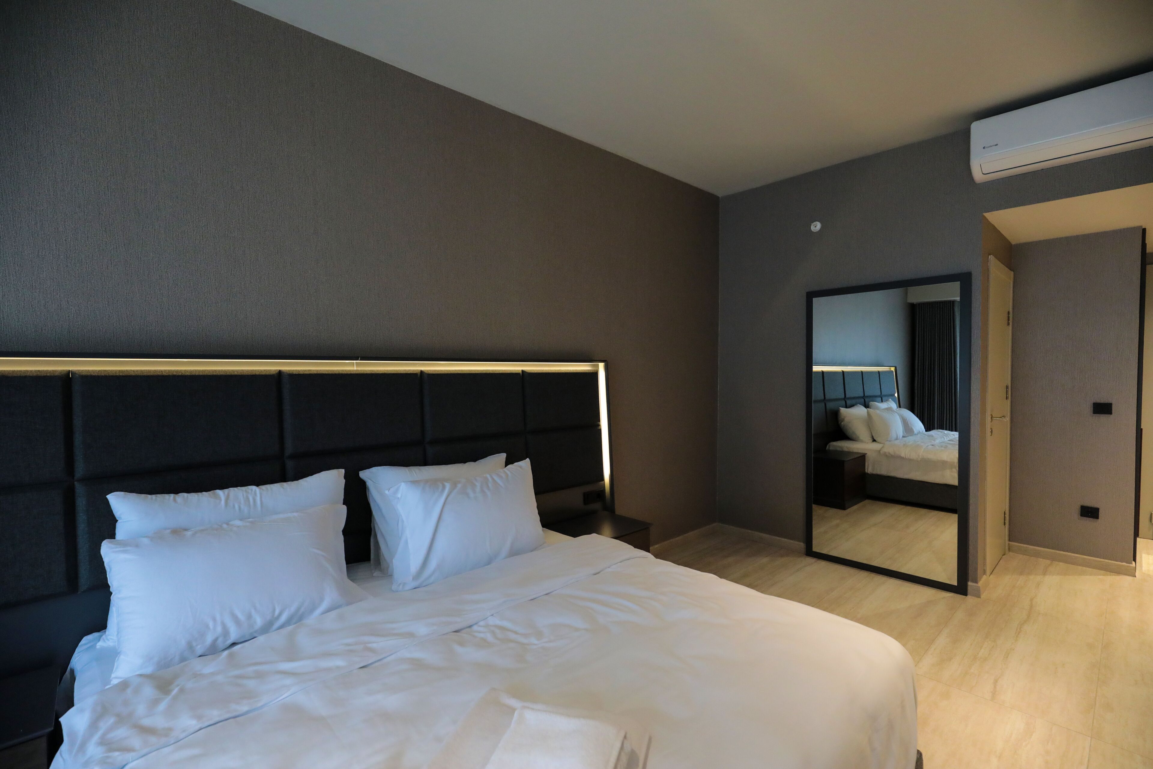 A modern hotel room with a large bed, a headboard, a bedside table, a mirror, and an air conditioning unit.