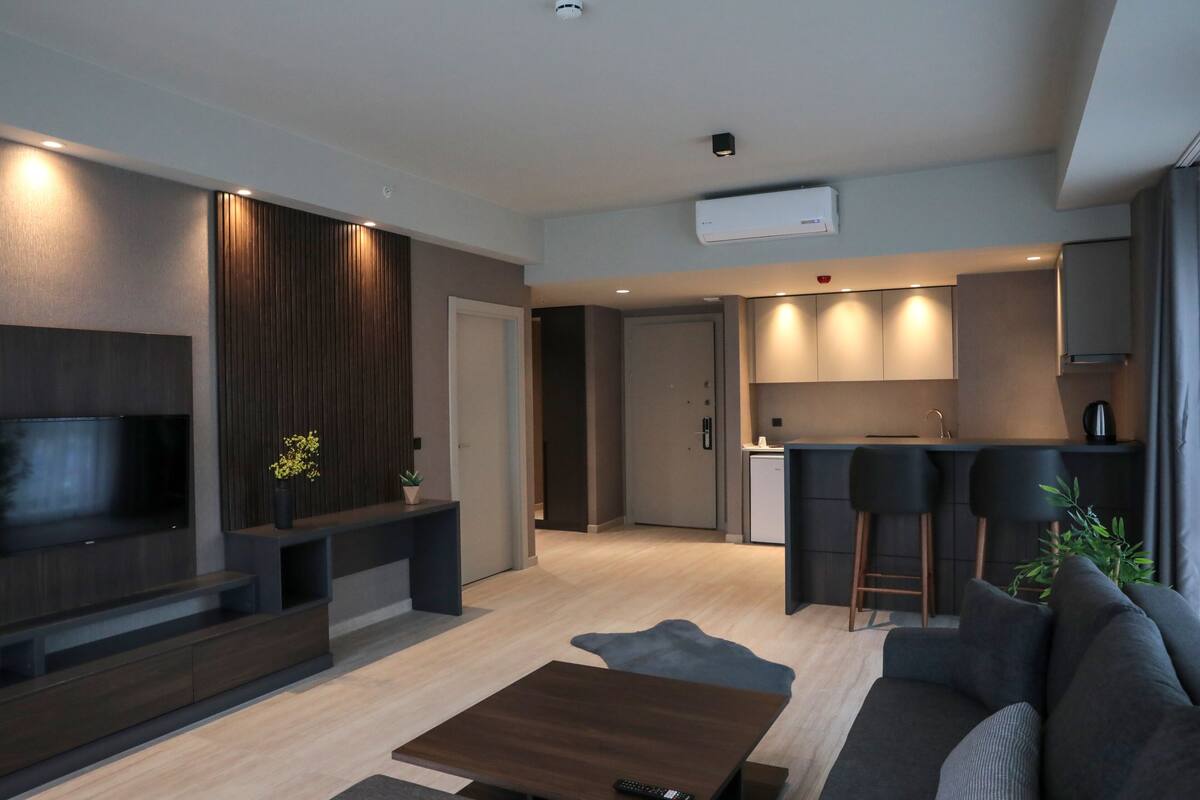A modern living room with a flat-screen TV, dark wood paneling, a small coffee table, and a gray sofa. A kitchen with bar stools and a countertop is visible in the background.