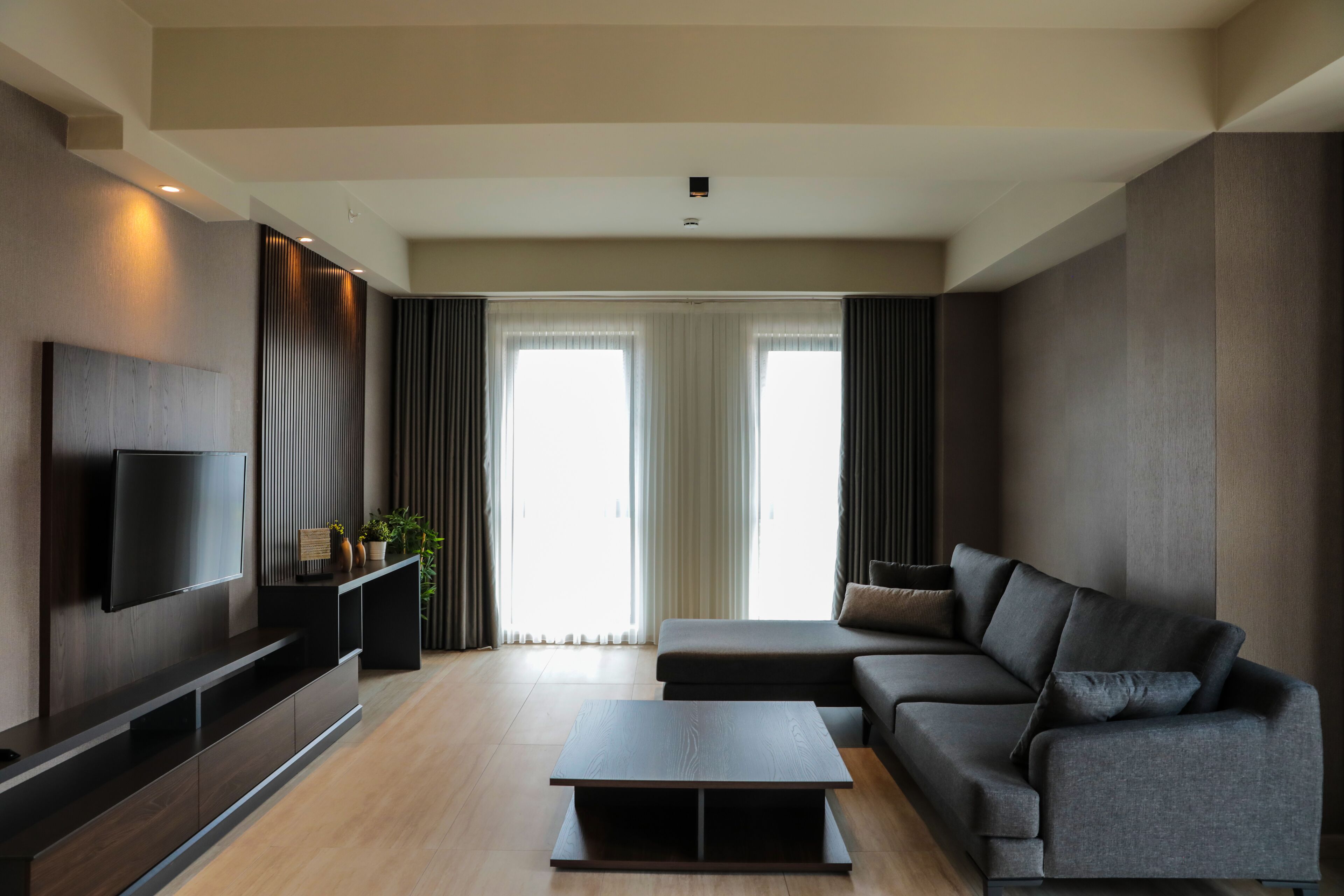 A modern living room with a flat-screen TV mounted on a dark wooden wall, a grey sofa, a coffee table, and a large window with curtains.