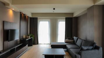 A modern living room with a flat-screen TV mounted on a dark wooden wall, a grey sofa, a coffee table, and a large window with curtains.