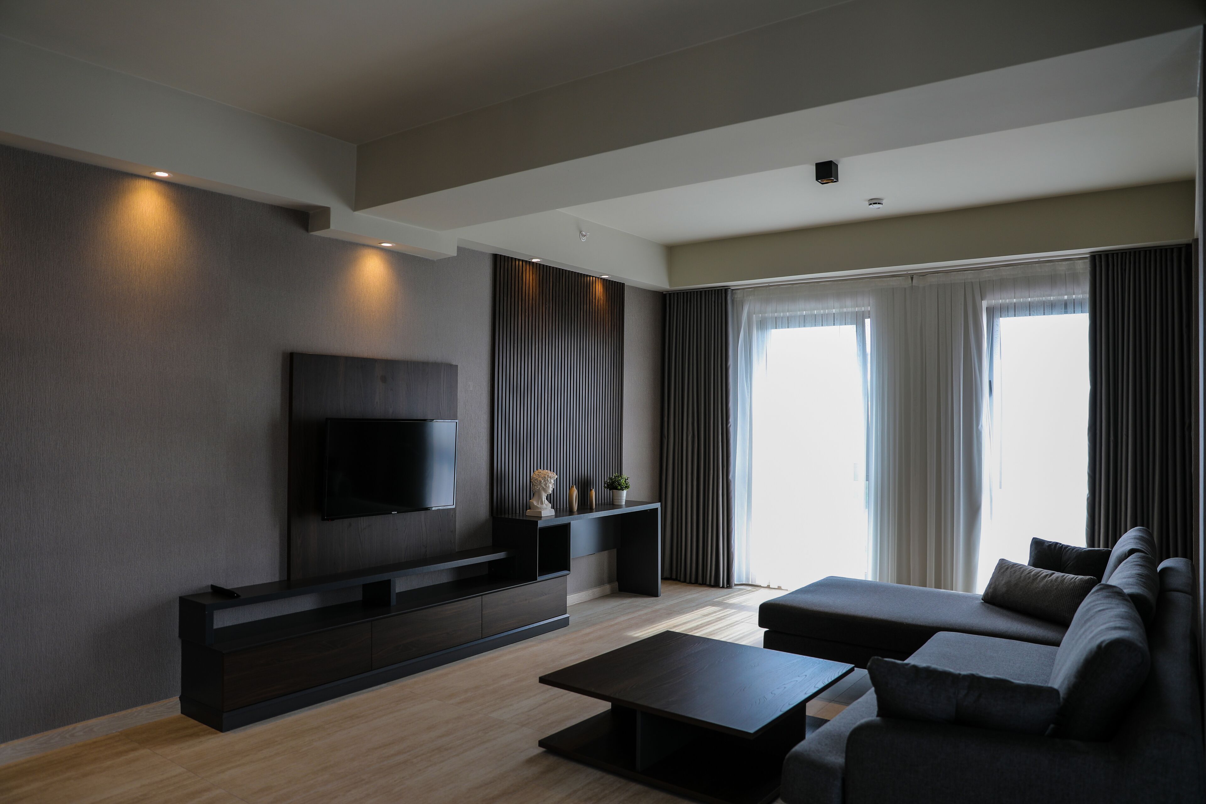 A modern living room with a flat-screen TV, dark furniture, and a large window with curtains.