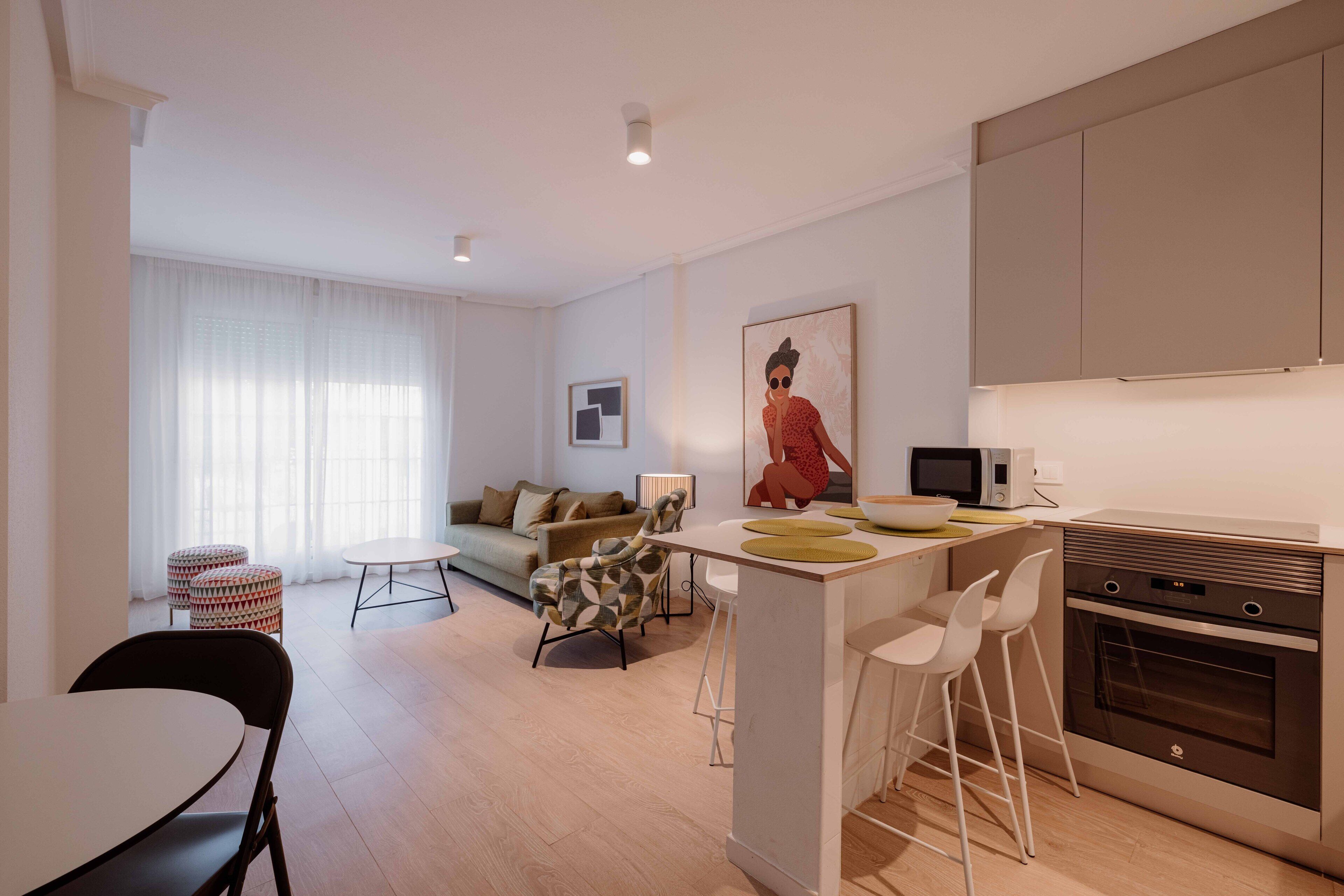 Una cocina moderna con una isla central, taburetes de bar, microondas y horno. Hay una mesa de comedor con sillas, un sofá y una ventana con cortinas de gasa.