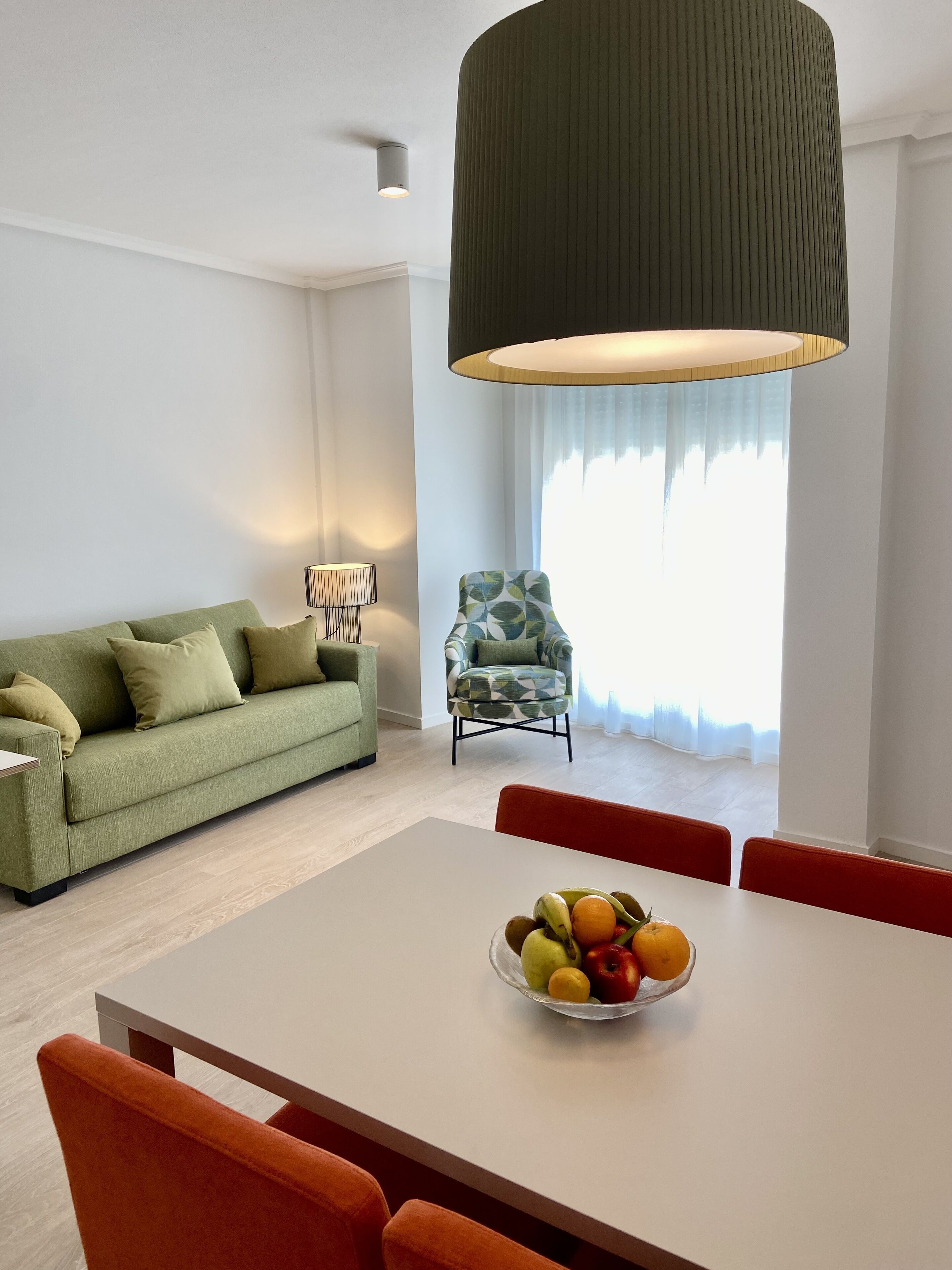 A modern living room with a green sofa, a patterned armchair, a dining table with a fruit bowl, and a large pendant light.