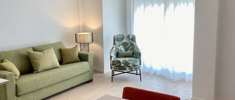 Ein modernes Wohnzimmer mit einem grünen Sofa, einem gemusterten Sessel, einem Esstisch mit Obstschale und einer großen Pendelleuchte.
