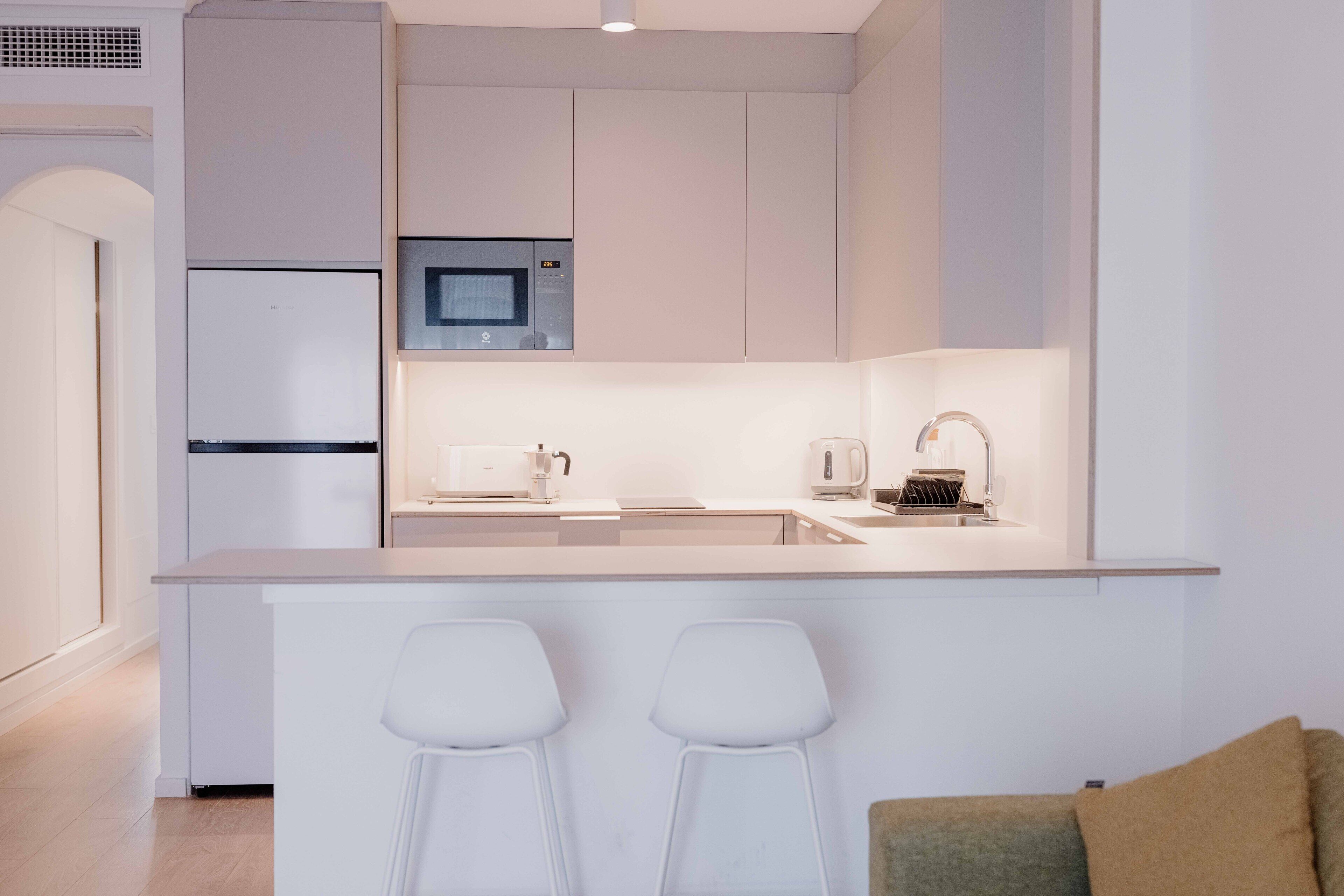 A modern kitchen with a built-in microwave, a refrigerator, and a coffee machine. There are two white bar stools in front of the counter.