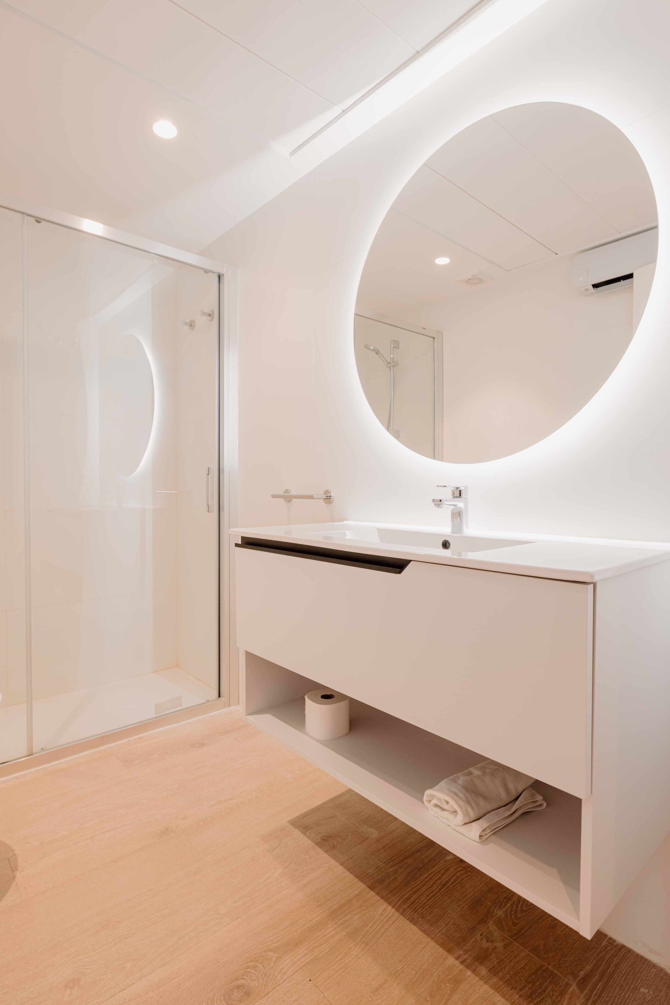 A modern bathroom with a white vanity, a round mirror, and a glass shower enclosure.