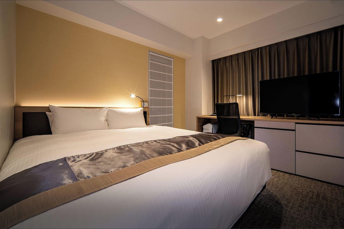 Ein Hotelzimmer mit einem großen Bett, einem Schreibtisch, einem Flachbildfernseher, einem Sessel und einem Fenster mit Vorhängen.