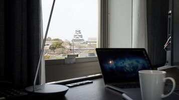 A laptop on a desk with a white mug, a remote control, and a window view of a building with a tower.