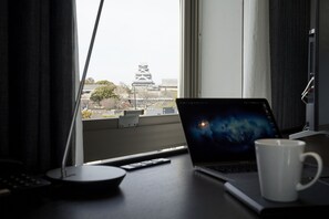 A laptop on a desk with a white mug, a remote control, and a window view of a building with a tower. - Daiwa Roynet Hotel Kumamoto (Kumamoto)