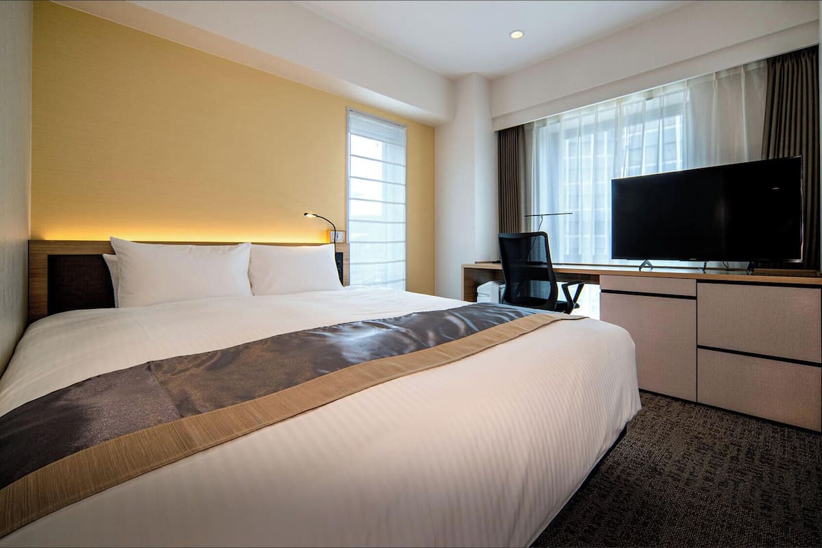 Ein Hotelzimmer mit einem großen Bett, einem Schreibtisch mit Stuhl, einem Flachbildfernseher und einem Fenster mit Vorhängen.