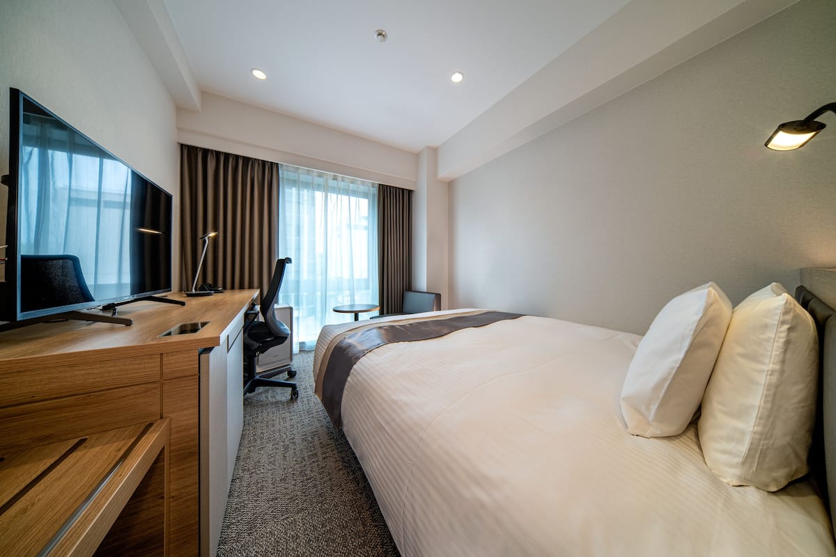 Ein Hotelzimmer mit einem Bett, einem Schreibtisch mit Computer, einem Sessel, einem Fernseher und einem Fenster mit Vorhängen.