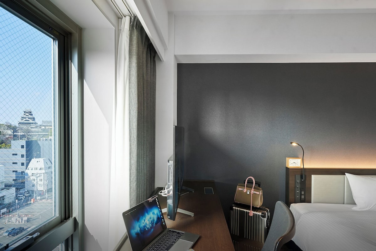 Ein Hotelzimmer mit großem Fenster, einem Schreibtisch mit Laptop, einem Bett, einem Nachttisch mit Lampe und Blick auf die Stadt.